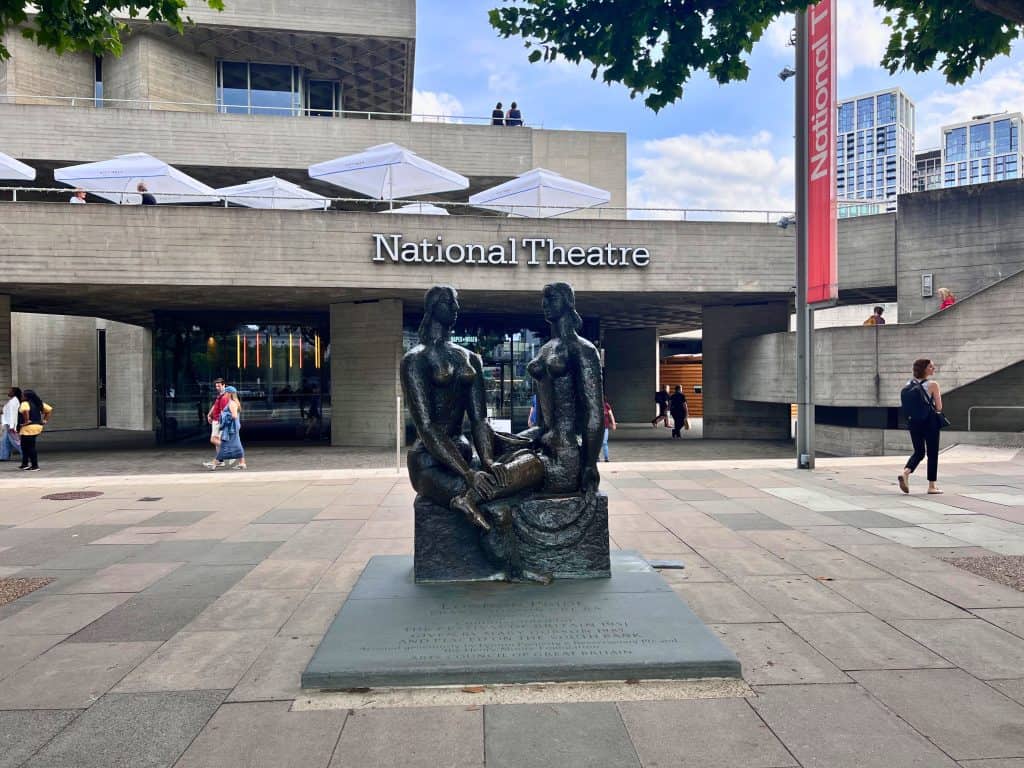 Concrete building with National Theatre written over the entrance. There is a black sculpture of two seated figures in front of the building.