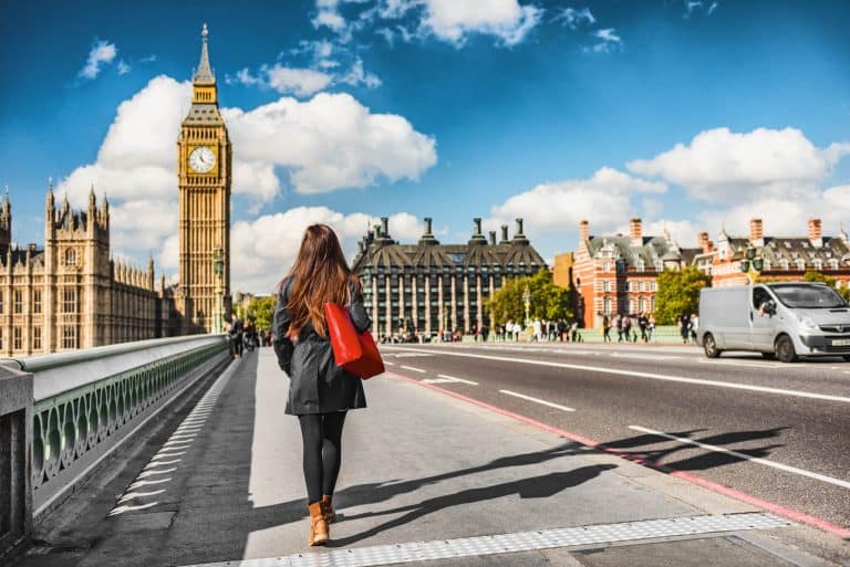A woman carrying a red bag, walking away from the camera across a bridge to the Houses of Parliament